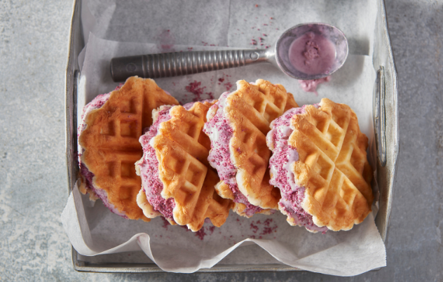 Four biscuit waffle ice cream sandwiches on parchment paper beside an ice cream scoop in a metal serving tray.